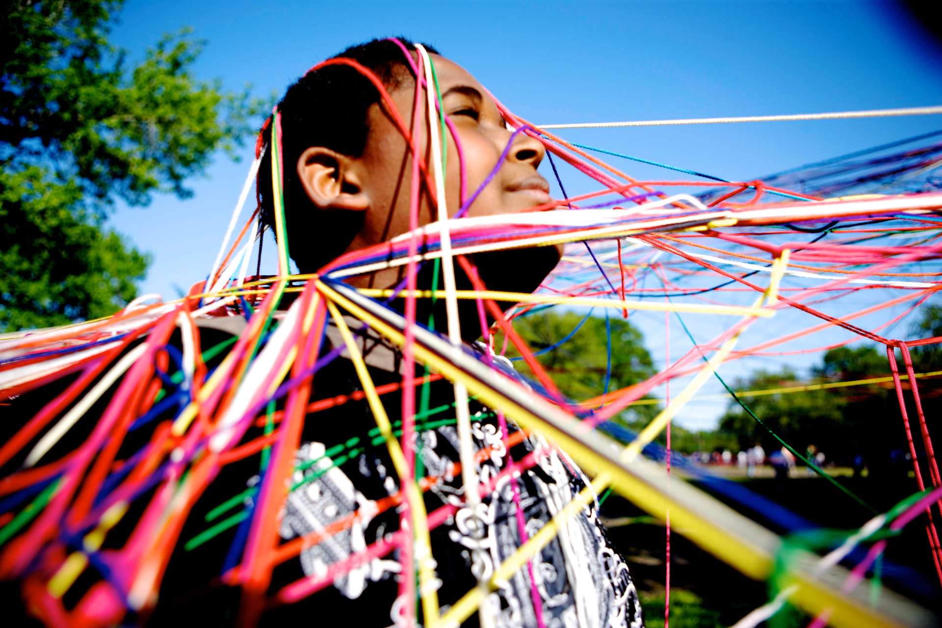 A child stands in the sun with their eyes closed, wrapped in a web of rainbow-colored strings from Jasmine Zimmerman's The Web Project installation